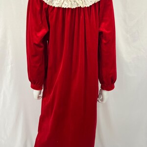 Op de afbeelding: Een lange, rode fluwelen nachtjapon met lange mouwen en een witte kanten kraag. De nachtjapon heeft een klassiek, elegant ontwerp. De kraag is afgewerkt met delicate kanten details.