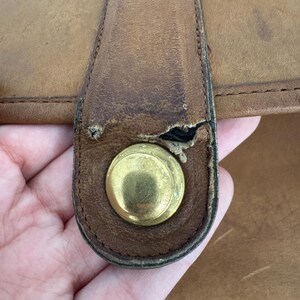 May include: Close-up of a tan leather bag with a brass-colored clasp. The leather shows signs of wear and tear, with visible scratches and a worn area near the clasp. The bag appears to be vintage or well-used.