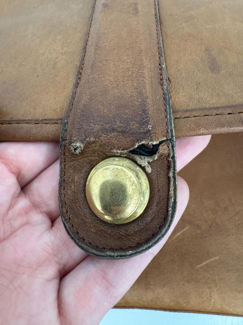 May include: Close-up of a tan leather bag with a brass-colored clasp. The leather shows signs of wear and tear, with visible scratches and a worn area near the clasp. The bag appears to be vintage or well-used.