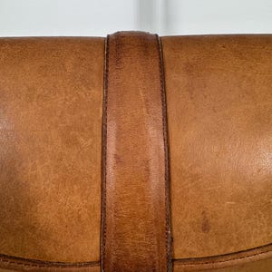 May include: Close-up of a vintage brown leather clutch with a strap closure. The leather has a worn, textured appearance with darker brown accents on the strap. A gold-toned clasp secures the flap.