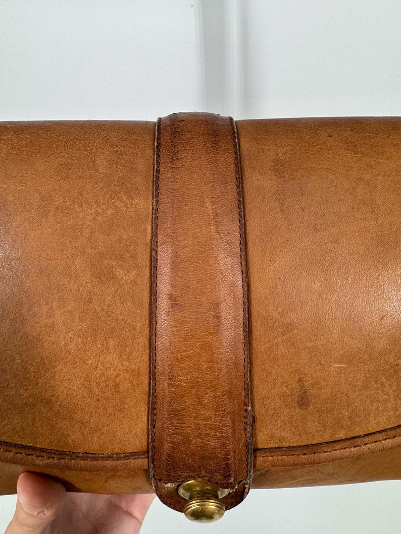 May include: Close-up of a vintage brown leather clutch with a strap closure. The leather has a worn, textured appearance with darker brown accents on the strap. A gold-toned clasp secures the flap.