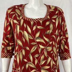 May include: A red dress with a square neckline and three-quarter sleeves. The dress features a repeating pattern of beige and green leaves and stems. The neckline and sleeve cuffs are trimmed with a striped pattern.