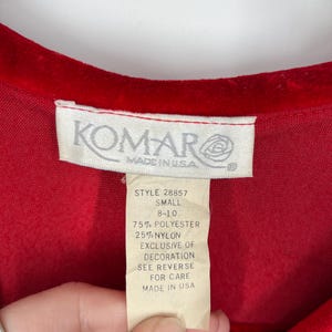 Op de afbeelding: Close-up van een rood kledingstuk met een label. Het label luidt "KOMAR MADE IN USA" en geeft het artikelnummer, de maat en de samenstelling van de stof weer: 75% polyester, 25% nylon. Het label bevat ook wasvoorschriften.