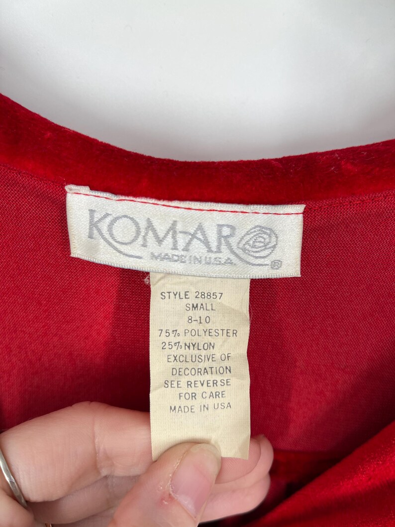 Op de afbeelding: Close-up van een rood kledingstuk met een label. Het label luidt "KOMAR MADE IN USA" en geeft het artikelnummer, de maat en de samenstelling van de stof weer: 75% polyester, 25% nylon. Het label bevat ook wasvoorschriften.
