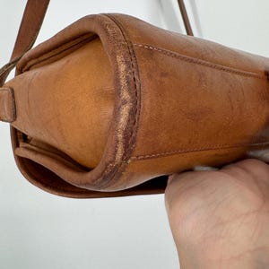 May include: A close-up of a tan leather crossbody bag. The bag has a rectangular shape with rounded edges and a long, thin strap. The leather shows signs of wear, with some discoloration and scuffing, giving it a vintage look.