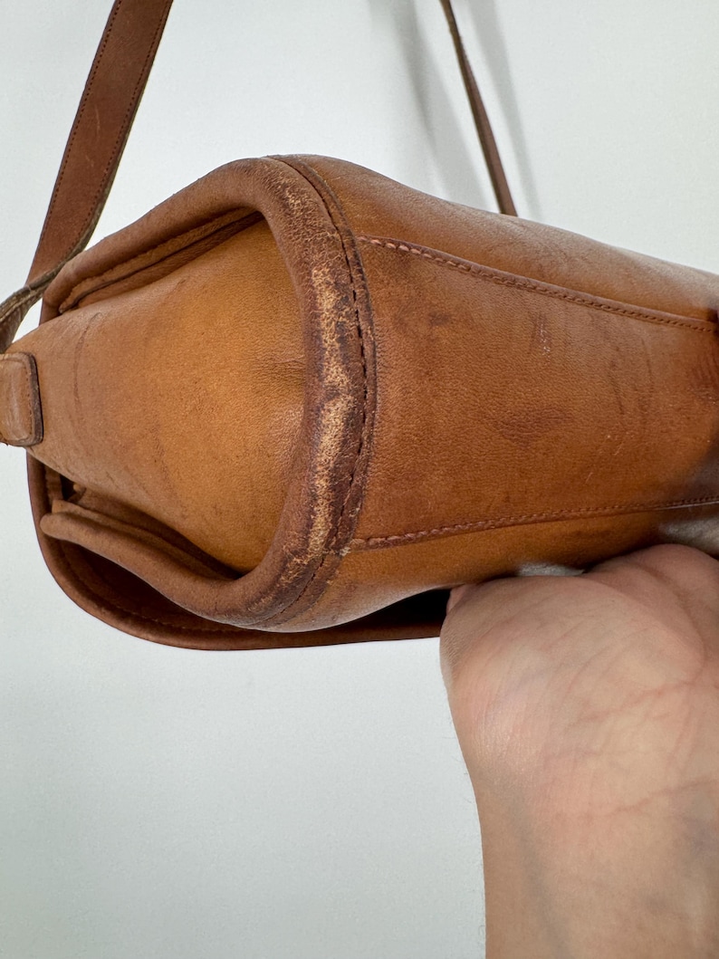 May include: A close-up of a tan leather crossbody bag. The bag has a rectangular shape with rounded edges and a long, thin strap. The leather shows signs of wear, with some discoloration and scuffing, giving it a vintage look.