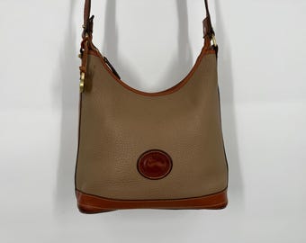 May include: A tan leather shoulder bag with a brown leather trim and a brown leather logo patch. The bag has a long strap and a flap closure.