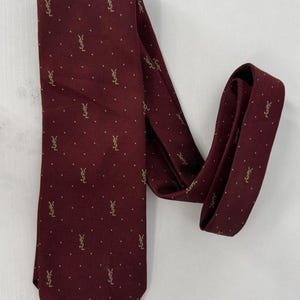 May include: A burgundy tie with a repeating pattern of small gold-colored logos and dots. The tie is folded over itself, showcasing the design. The tie is made of a smooth material.