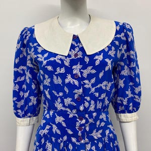 May include: A vibrant blue dress featuring a repeating white leaf pattern, a large cream-colored Peter Pan collar, and cream-colored sleeve cuffs. The dress has a front button-down closure with small purple buttons and three-quarter length sleeves.