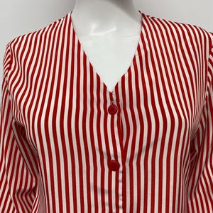 May include: A red and white striped blouse with a V-neckline and long sleeves. The blouse has red buttons down the front and is displayed on a mannequin.