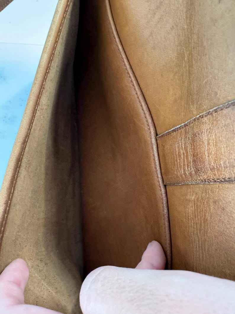 May include: Open view of a brown leather bag's interior. The bag's interior is a lighter shade of brown than the exterior. The stitching is visible along the seams. The bag appears to be well-worn.