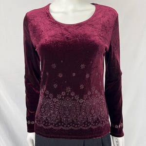 May include: A long-sleeved, burgundy velvet top with a scoop neck. The shirt features a detailed, embroidered paisley design along the bottom hem and cuffs. Small embroidered floral accents are scattered across the upper portion of the shirt.