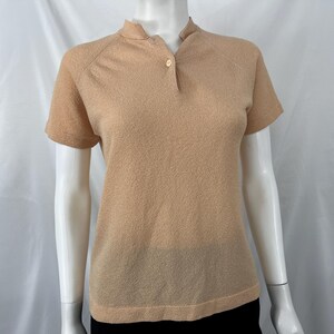 May include: A short-sleeved, tan-colored knit shirt with a mandarin collar and a single button closure. The shirt has a textured appearance and a classic, simple design, suitable for casual wear.