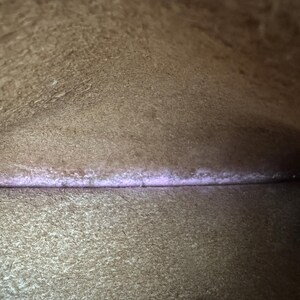 May include: Interior view of a brown leather bag. The image shows the inside of the bag, with a visible seam and a subtle purple hue along the seam. The leather has a textured appearance.