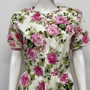 May include: A white dress with a pink rose floral pattern. The dress has short sleeves, a round neckline, and a cinched waist. The neckline features a decorative tie closure.