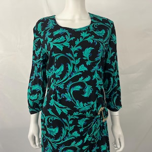 May include: A black dress featuring a vibrant teal and blue swirling leaf pattern, a round neckline, and three-quarter length sleeves. The garment includes a gathered side detail secured with a circular gold-toned metal accent at the waist.