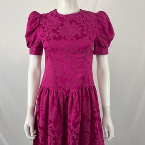 May include: A magenta-colored vintage dress with a floral pattern. The dress features short, puffed sleeves and a fitted bodice that flares into a skirt. The dress is displayed on a mannequin.