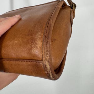 May include: A vintage brown leather crossbody bag with a long strap. The bag has a rectangular shape with rounded corners and visible stitching. The leather shows signs of wear, adding to its character.