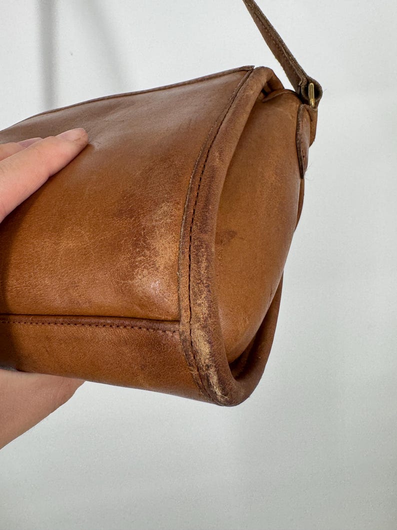 May include: A vintage brown leather crossbody bag with a long strap. The bag has a rectangular shape with rounded corners and visible stitching. The leather shows signs of wear, adding to its character.