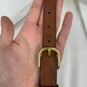 May include: Close-up of a brown leather strap with a gold-colored buckle. The strap has several adjustment holes and is likely part of a bag or accessory. The leather shows signs of wear.