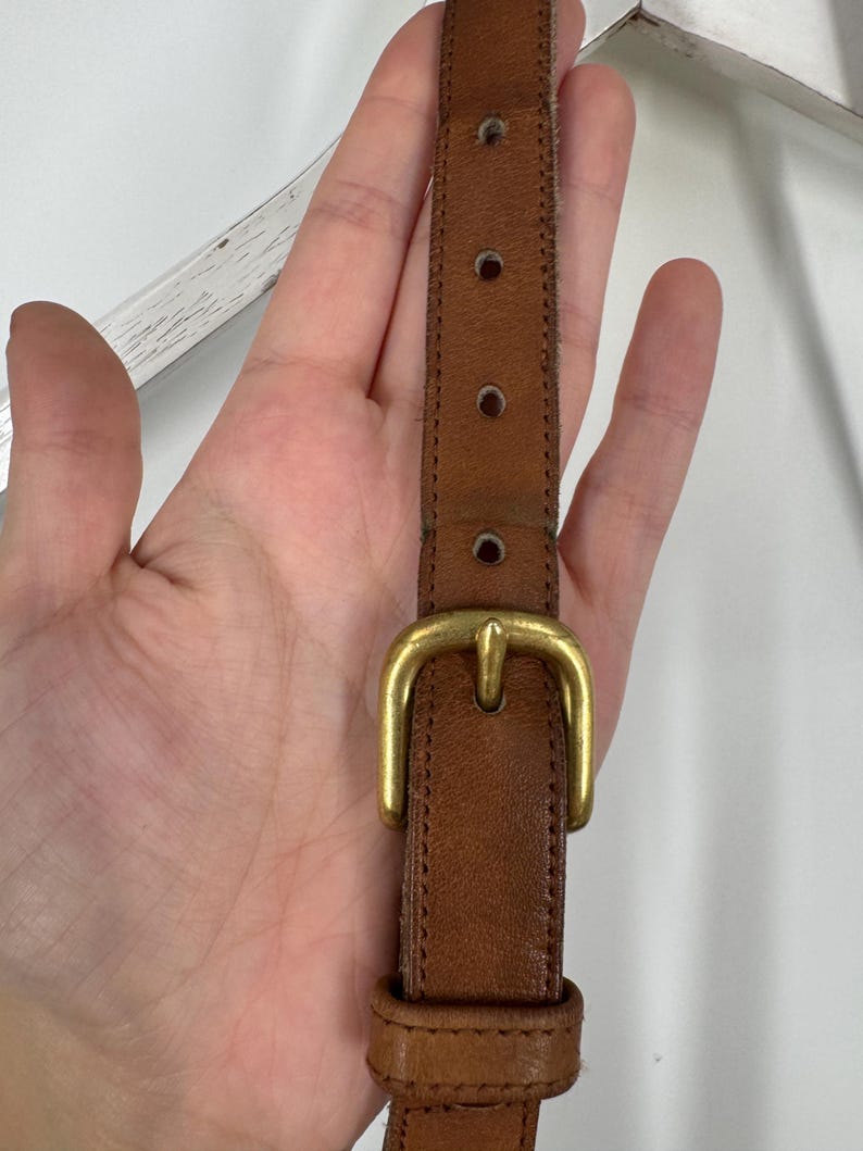 May include: Close-up of a brown leather strap with a gold-colored buckle. The strap has several adjustment holes and is likely part of a bag or accessory. The leather shows signs of wear.