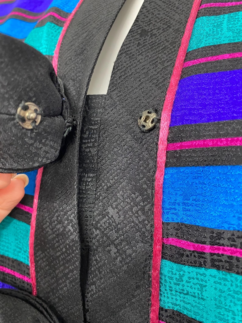 May include: Close-up of a vintage jacket with a black textured lapel and a colorful striped pattern. The stripes include teal, blue, purple, and pink. The jacket has a black button closure.