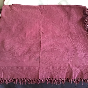 May include: A burgundy-colored textile with an embossed design featuring circular motifs and floral patterns. The textile has a fringed edge and is likely a throw or decorative blanket. The overall aesthetic is vintage or antique.