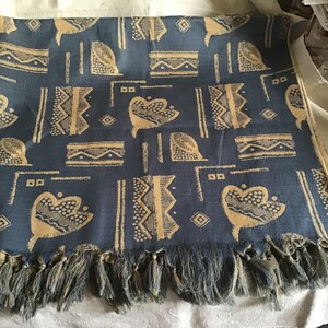 May include: A blue textile with a repeating pattern of beige abstract shapes and lines. The fabric has a fringe along the bottom edge. This decorative textile could be used as a scarf, shawl, or throw.