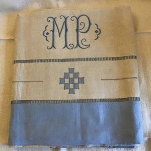 May include: A cream-colored linen tablecloth with blue embroidered details. The tablecloth features a monogram "MP" in the top center and a blue checkered design in the center. The bottom edge of the tablecloth is a solid blue.