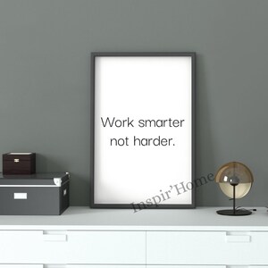 Puede incluir: Un cuadro de pared rectangular vertical en un marco negro fino con el texto negro Work smarter not harder. sobre un fondo blanco. La lámina se muestra sobre una superficie blanca junto a una caja de almacenamiento gris oscuro y una lámpara de cristal.