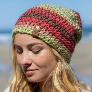 May include: A slouchy crochet beanie featuring a variegated yarn pattern in shades of green, brown, and pink. The hat includes a small, rectangular tan leather tag attached to the brim, embossed with a simple line-art heart design and a metal rivet.