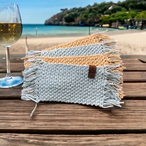 May include: Crochet coasters in light blue and tan with fringing. A leather tag with a coffee cup logo is attached. A glass of white wine sits next to the coasters on a wooden table, with a beach scene in the background. Coasters are ideal for drinks.
