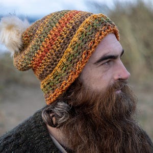 May include: A hand-crocheted beanie hat in autumnal colours of orange, green, and brown, featuring a cream-coloured pom-pom. The hat has a ribbed brim. Ideal for keeping warm in colder weather.