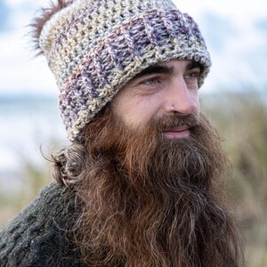 May include: A crocheted beanie in shades of beige, purple, and brown, with a brown faux fur pom-pom. The hat has a textured stitch pattern, suitable for cold weather. The beanie is shown on a person.
