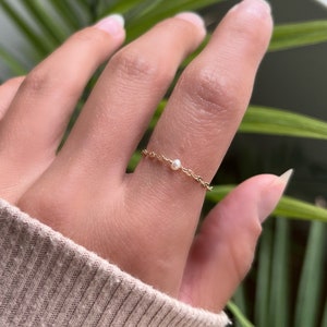 Puede incluir: Un delicado anillo de cadena de oro con un solo acento de perla. El anillo se lleva en un dedo con una manga de suéter marrón claro en el fondo.