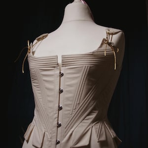May include: Cream-colored corset with a structured design, displayed on a mannequin. The bodice features vertical and horizontal stitching, a row of silver-toned buttons, and decorative shoulder straps. The corset has a fitted silhouette and a flared peplum.