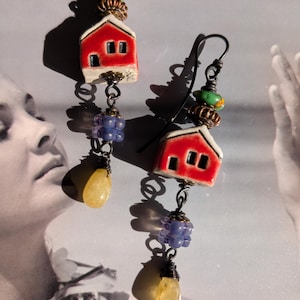 May include: A pair of drop earrings featuring miniature red ceramic house charms with white roofs. Each earring includes a green bead, a copper-toned spacer, a purple textured bead, and a yellow teardrop-shaped stone suspended on a dark metal wire.