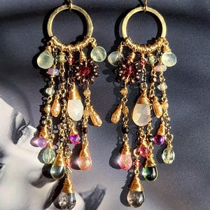 May include: A pair of gold earrings with a circular top and dangling strands of colourful beads and gemstones. The earrings feature a variety of colours, including green, pink, purple, and clear, with a mix of shapes and sizes. The earrings are designed for pierced ears.