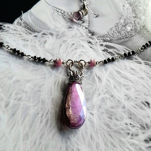 Op de afbeelding: Een zilveren ketting met een grote, gefacetteerde, paarse edelsteen hanger. De hanger is gezet in een zilveren kroonvormige setting. De ketting is versierd met kleine roze en zwarte kralen.