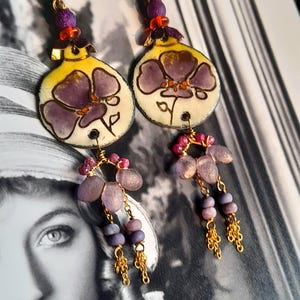 Op de afbeelding: Paarse bloemeoorbellen met gouden kettingen en paarse kralen. De oorbellen hebben een gele achtergrond met een paars bloemenontwerp.