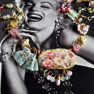 May include: A handmade necklace with a floral pendant displaying pink and white roses. The necklace incorporates various colourful beads, including pink, green, and yellow, strung on gold-coloured wire. A light blue fabric accent adds a soft touch.