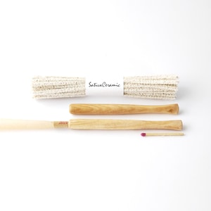 May include: An overhead shot shows a bundle of white pipe cleaners with a "Sativa Ceramic" label, two wooden tools, and a matchstick with a pink head. The tools are light brown with a handle shape.
