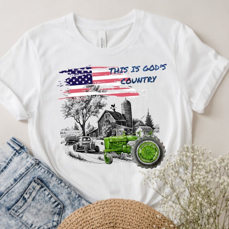 Puede incluir: Camiseta blanca con un dise&ntilde;o de bandera estadounidense desgastada y un tractor verde en una escena de granja. El texto "This is God's Country" est&aacute; impreso en la camiseta.