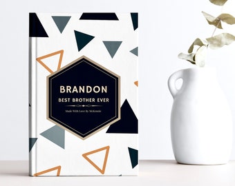 May include: A white notebook with a geometric pattern of black and gray triangles and orange triangles. The notebook has a black hexagon with gold trim and the text "BRANDON BEST BROTHER EVER Made With Love By McKenzie".