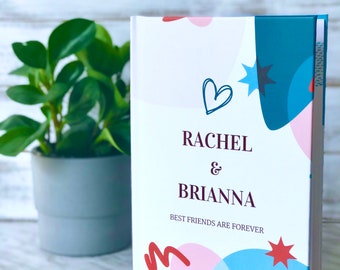 Op de afbeelding: Een wit hardback dagboek met een kleurrijk abstract ontwerp. De omslag heeft de namen "Rachel & Brianna" en de tekst "Best Friends Are Forever".