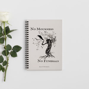 K&ouml;nnte beinhalten: Ein spiralgebundenes Notizbuch mit einem beigefarbenen Einband, der eine Schwarzwei&szlig;illustration eines Baumes mit einem Raben auf einem Ast zeigt. Der Text "No Mourners No Funerals Alice's Notebook" ist auf dem Einband gedruckt.