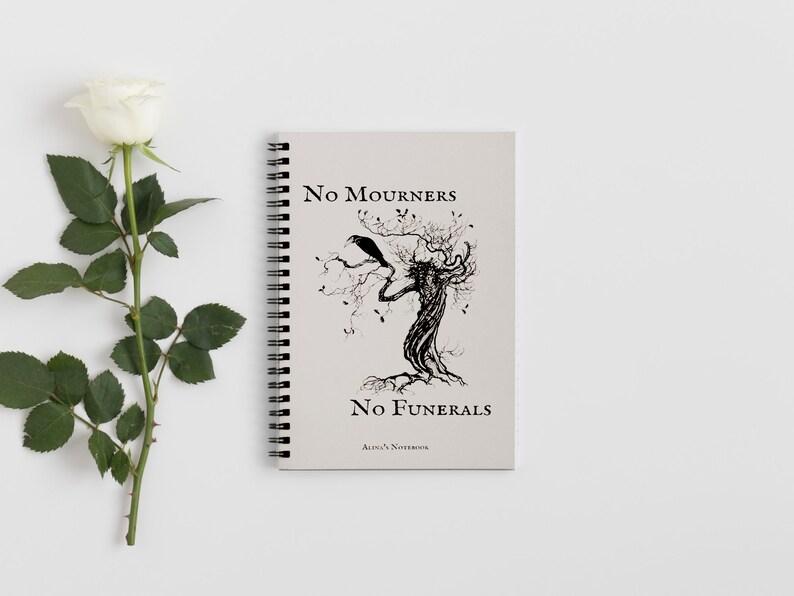 K&ouml;nnte beinhalten: Ein spiralgebundenes Notizbuch mit einem beigefarbenen Einband, der eine Schwarzwei&szlig;illustration eines Baumes mit einem Raben auf einem Ast zeigt. Der Text "No Mourners No Funerals Alice's Notebook" ist auf dem Einband gedruckt.