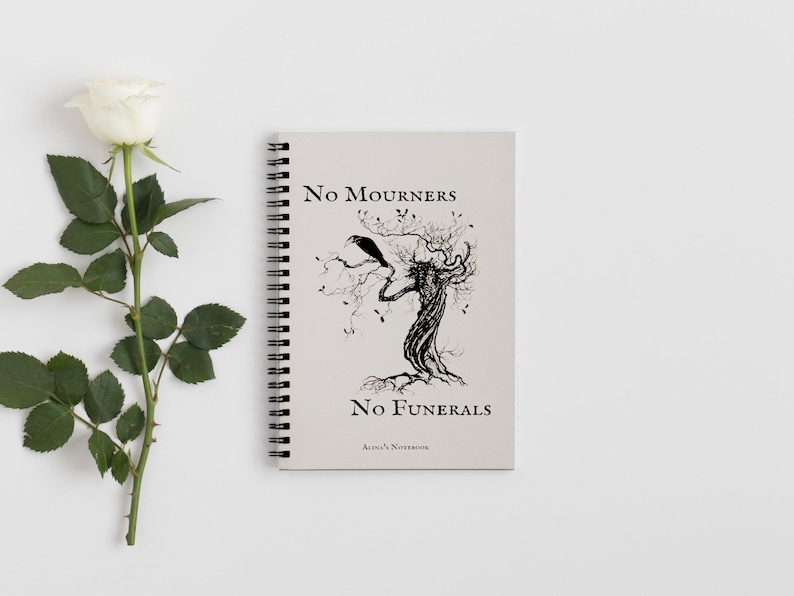 K&ouml;nnte beinhalten: Ein spiralgebundenes Notizbuch mit einer Schwarzwei&szlig;illustration eines Baumes mit einem Raben auf einem Ast. Der Text auf dem Cover lautet "No Mourners No Funerals Alice's Notebook".