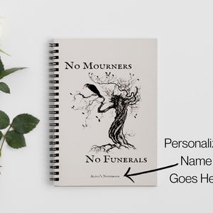 K&ouml;nnte beinhalten: Ein spiralgebundenes Notizbuch mit einer Schwarzwei&szlig;illustration eines Baumes mit einem Raben auf einem Ast. Der Text "No Mourners No Funerals" ist auf dem Cover gedruckt. Das Notizbuch liegt auf einem wei&szlig;en Hintergrund.