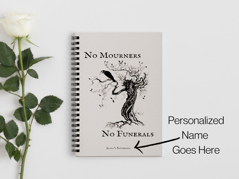 K&ouml;nnte beinhalten: Ein spiralgebundenes Notizbuch mit einer Schwarzwei&szlig;illustration eines Baumes mit einem Raben auf einem Ast. Der Text "No Mourners No Funerals" ist auf dem Cover gedruckt. Das Notizbuch liegt auf einem wei&szlig;en Hintergrund.
